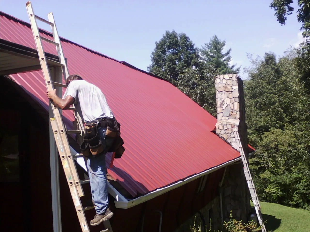 Roofer on a ladder beside a red metal roof and stone chimney in Industry